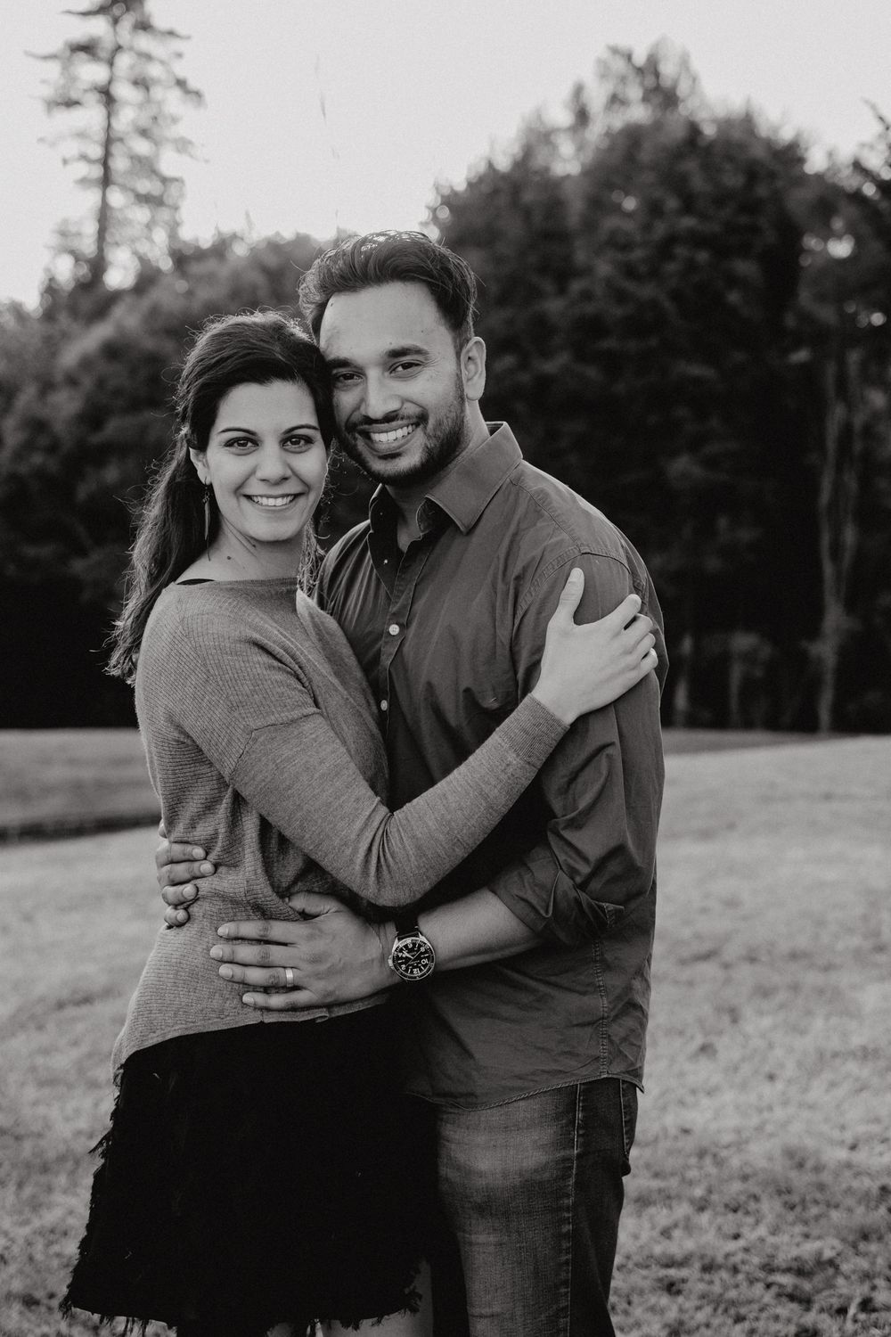 A couple embraces while posing outdoors in matching casual outfits with trees in the background.