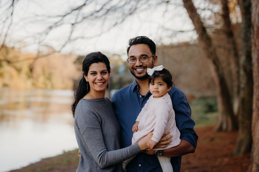 A family stands together smiling by a lake during autumn with tall trees in the background.