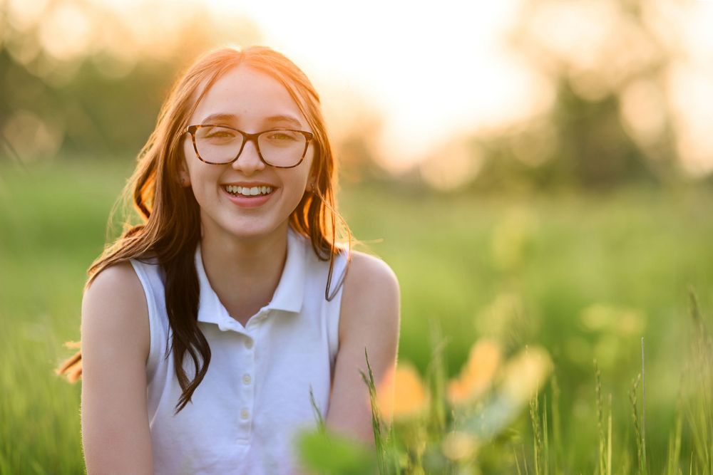 A young person with glasses smiling in a sunny meadow at golden hour, wearing a white sleeveless top.
