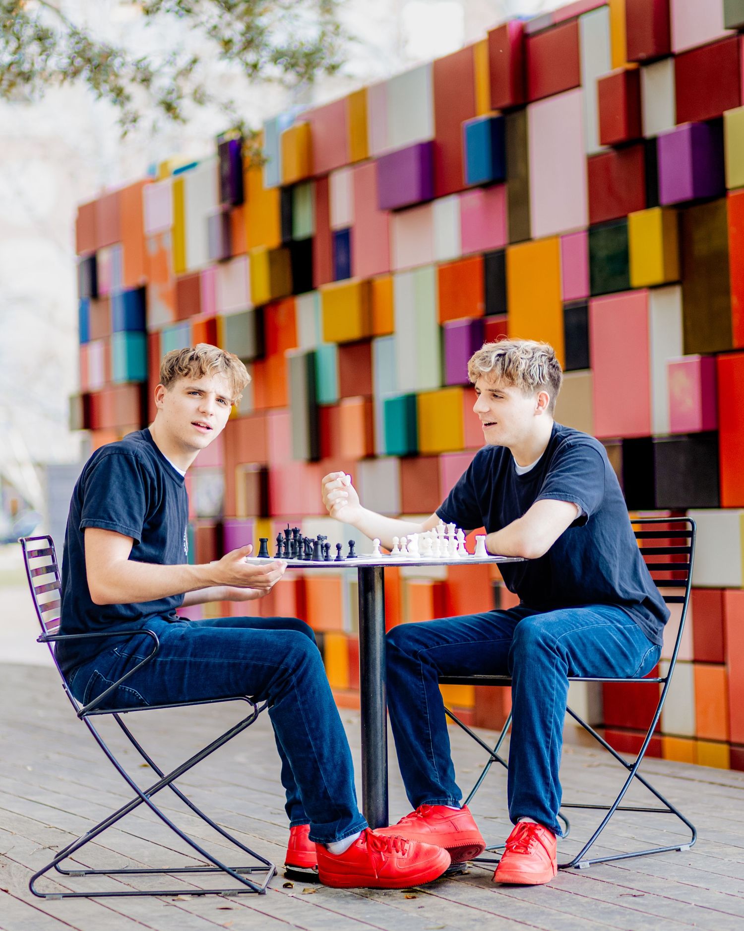 Two people playing chess at a table outdoors against a colorful geometric wall backdrop.