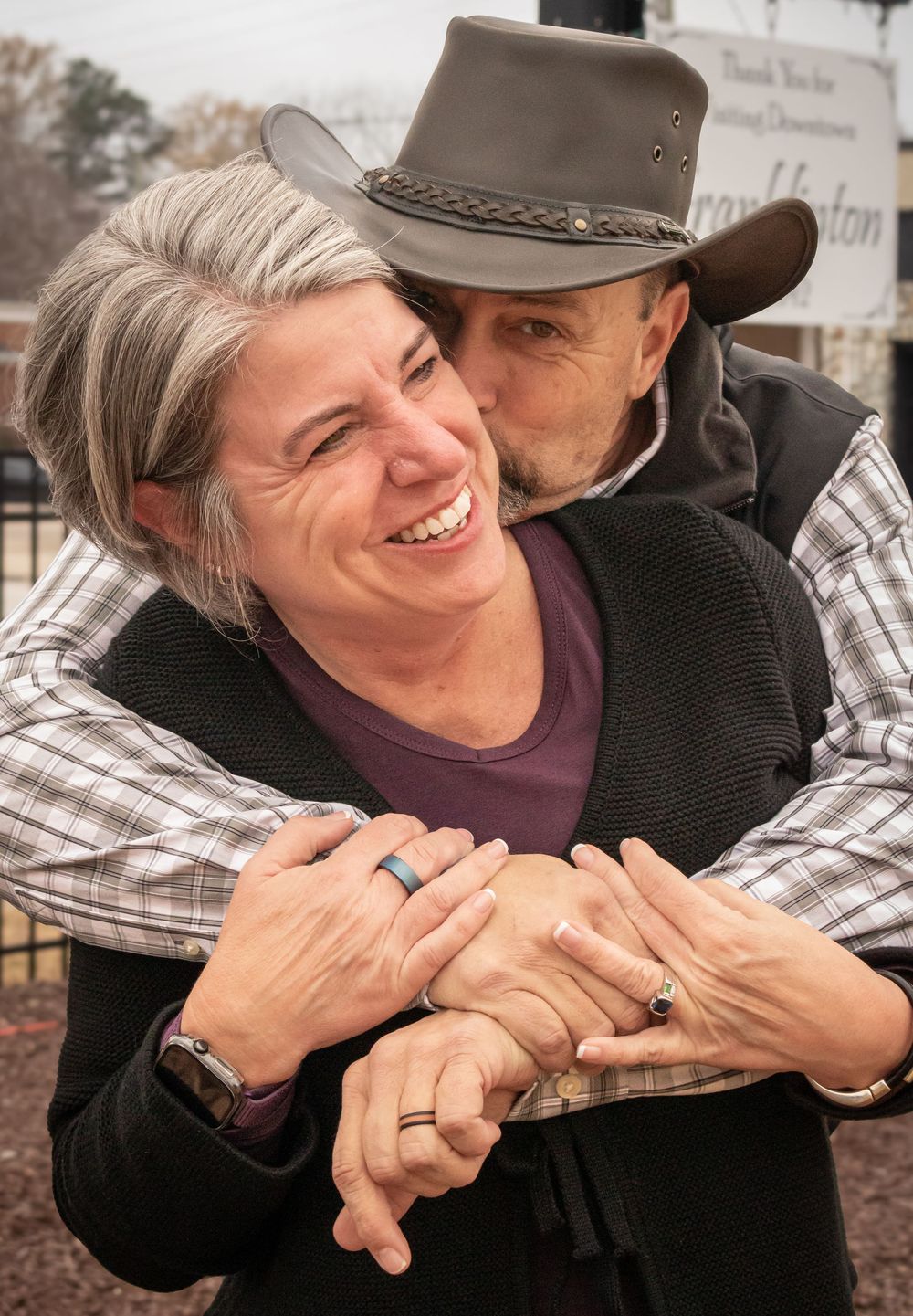 A couple shares a tender moment outdoors, with one wearing a cowboy hat and plaid shirt embracing the other from behind.