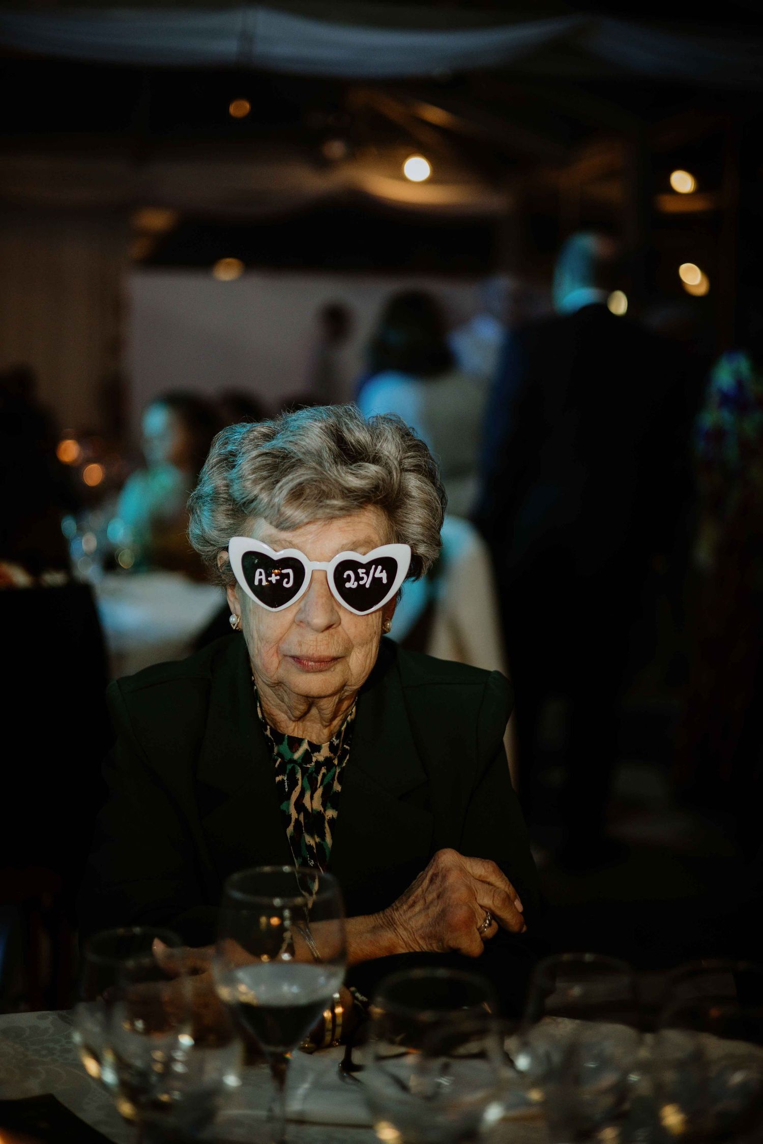 An elderly person wearing reflective sunglasses sits at a dimly lit table during an evening event.
