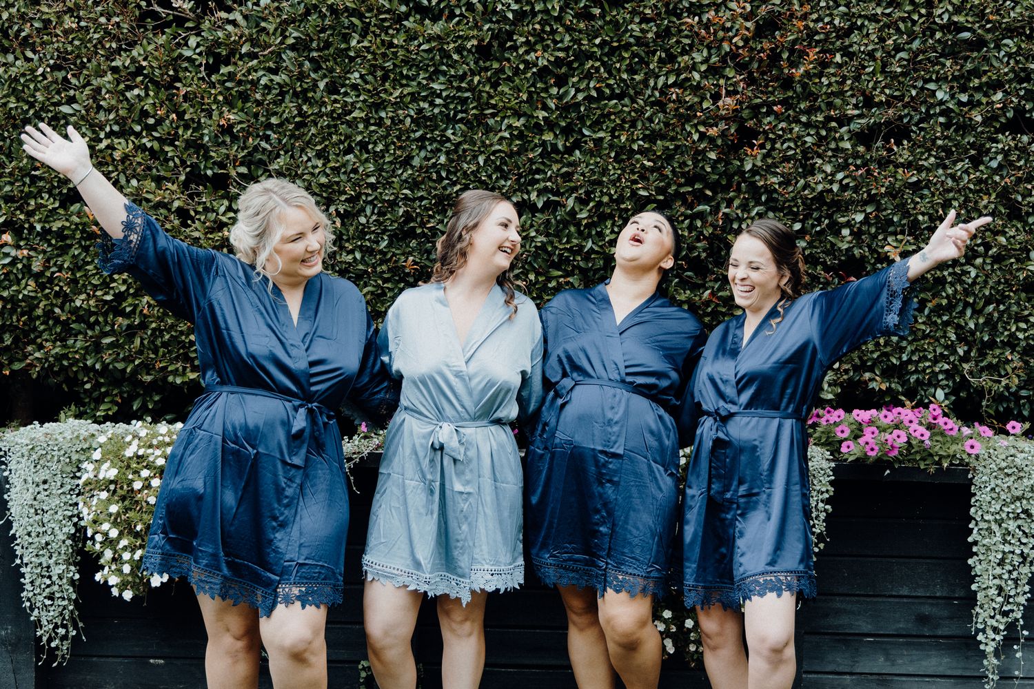 Girl squad in a blue silk robes poses joyfully in a garden setting, captured by Apertured Media Auckland Wedding Photographer.