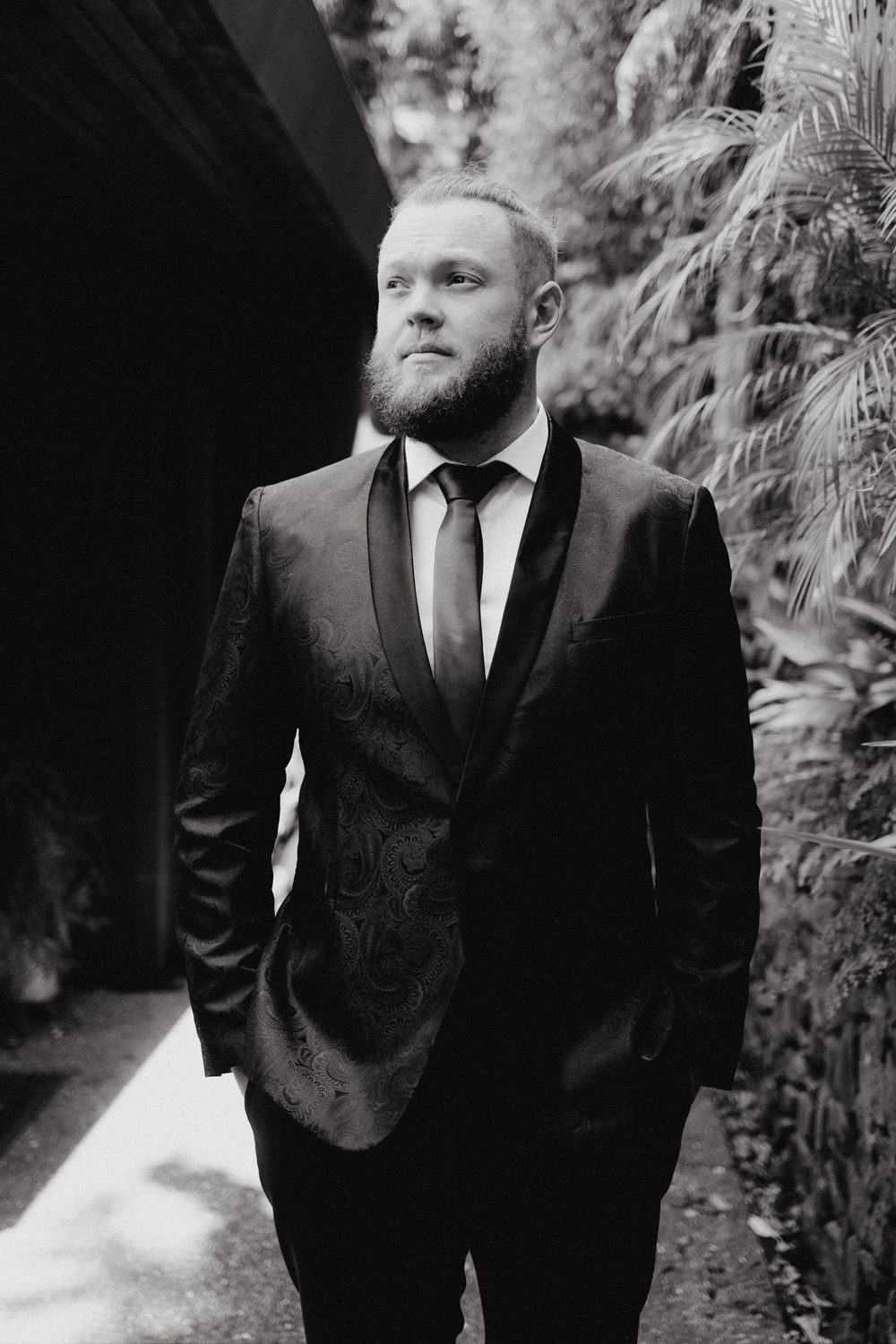 Black and white portrait of the groom standing outdoors near lush foliage, captured by Apertured Media Auckland Wedding Photographer.