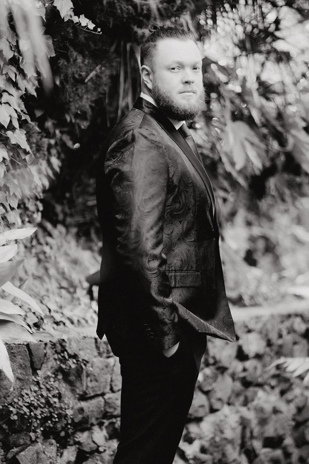 The Groom in a formal suit jacket posing against a leafy outdoor backdrop, captured by Apertured Media Auckland Wedding Photographer.