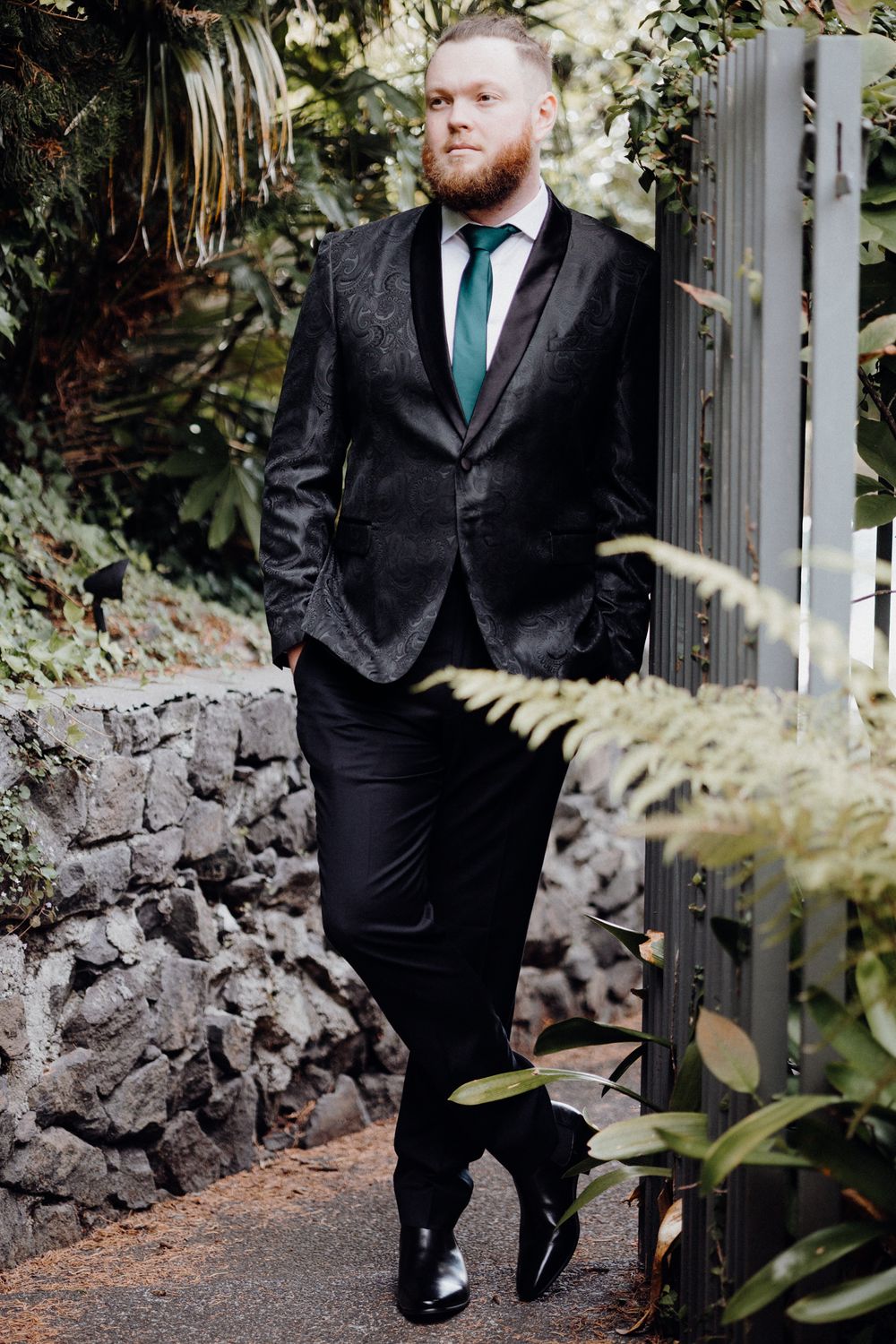 The groom with emerald green tie poses against stone wall , captured by Apertured Media Auckland Wedding Photographer.
