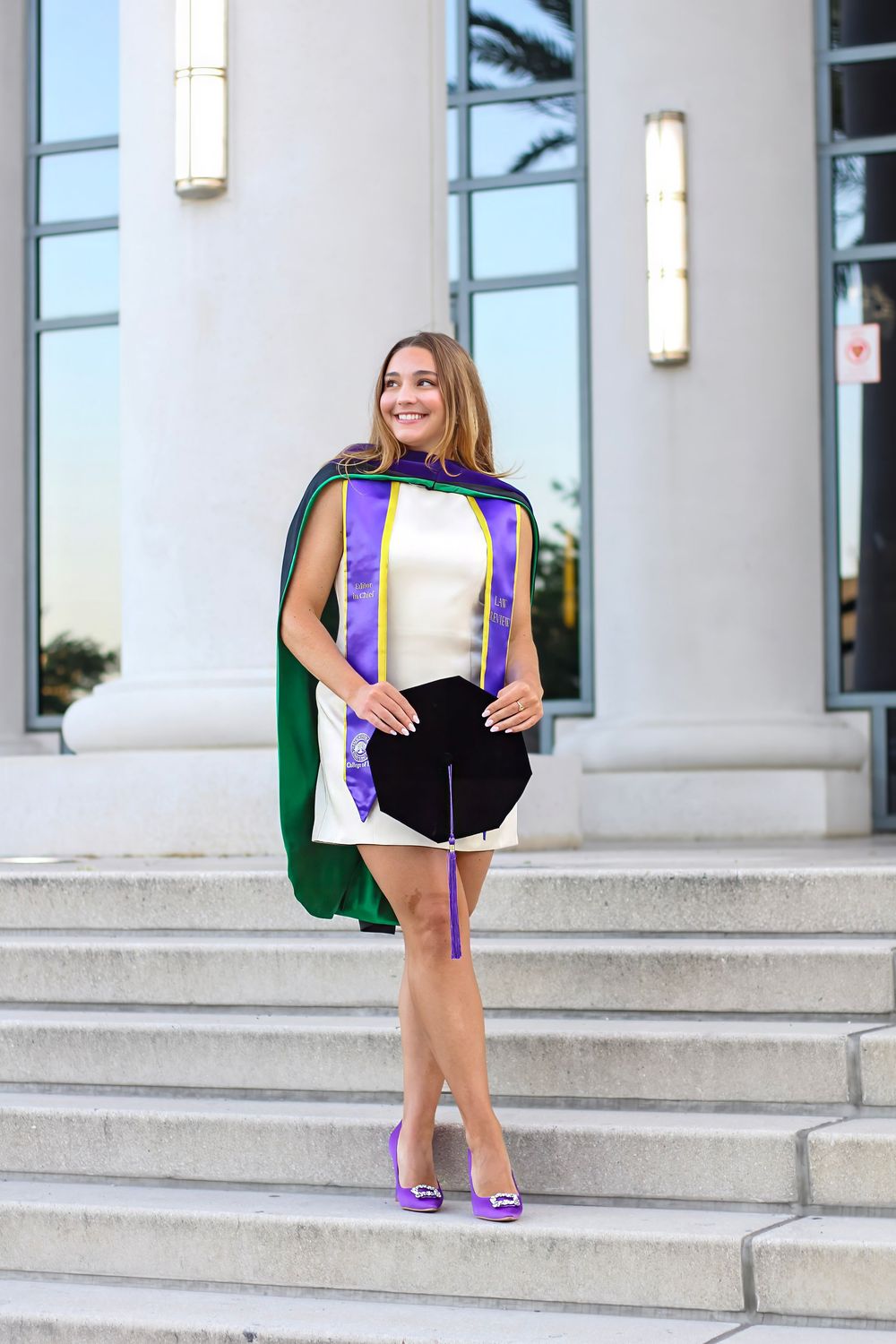 Graduate in purple and black regalia posing on concrete steps of academic building.