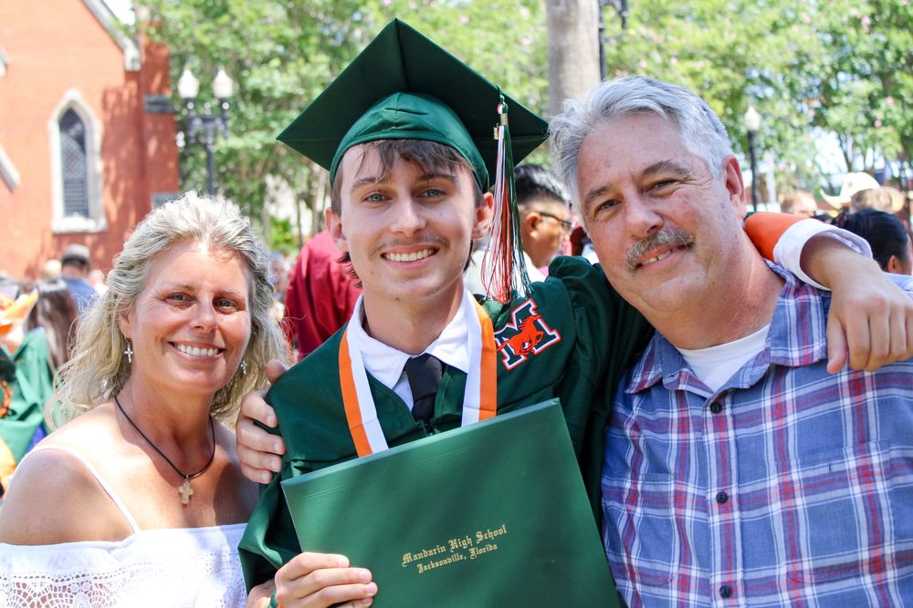 Graduates celebrating with diplomas in green caps and gowns during an outdoor ceremony.