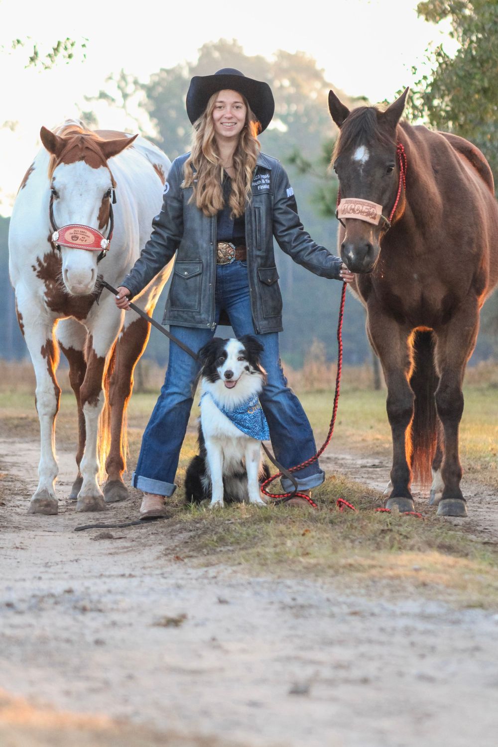 Person in denim and hat leading horses and dogs on a dirt path during golden hour.