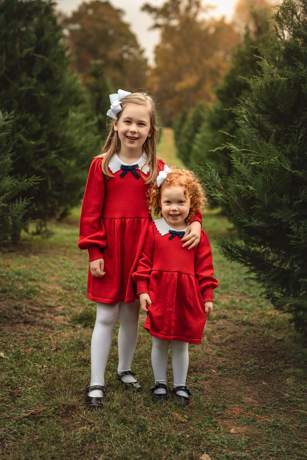 Two sisters in matching red dresses with white collars stand together among evergreen trees at sunset.
