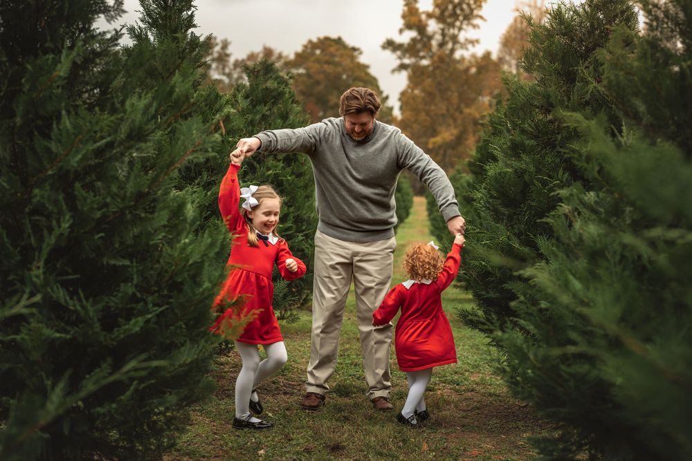 Dad twirls his young daughters in red as they dance between rows of Christmas trees for Christmas mini session