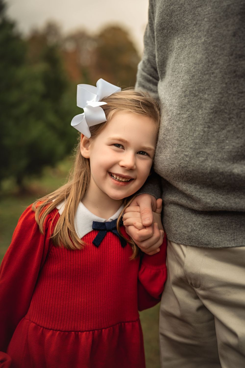 A girl in a red sweater dress and white bow create a cheerful holiday portrait as she holds her father's hand