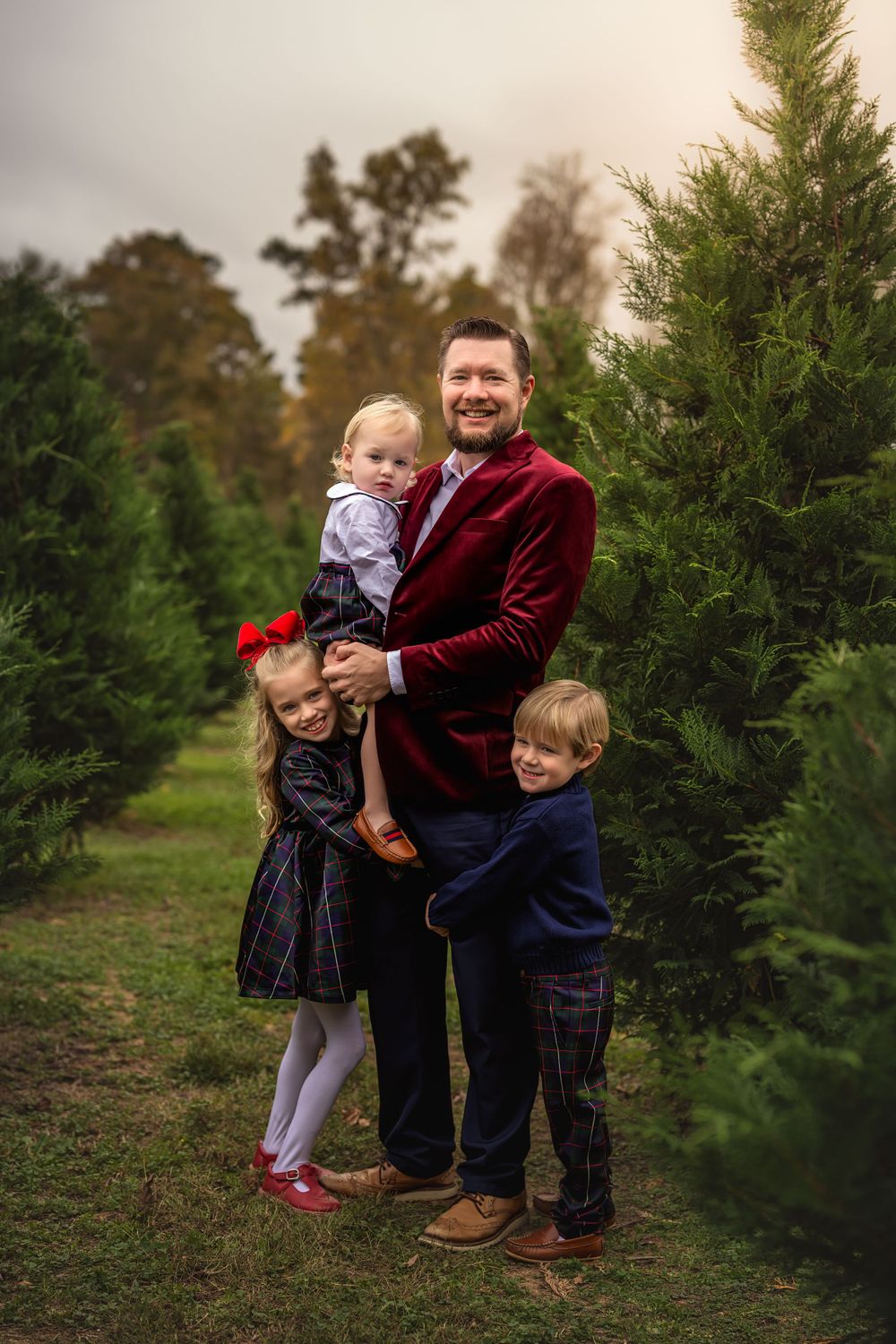 A well-dressed dad poses with his children for a holiday portrait among evergreen trees at a Christmas tree farm.
