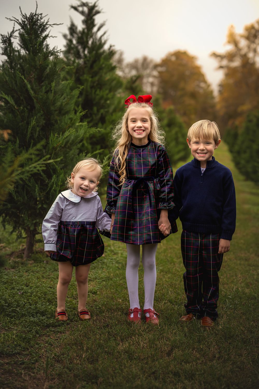 Three children in coordinating plaid outfits pose together at a Christmas tree farm.