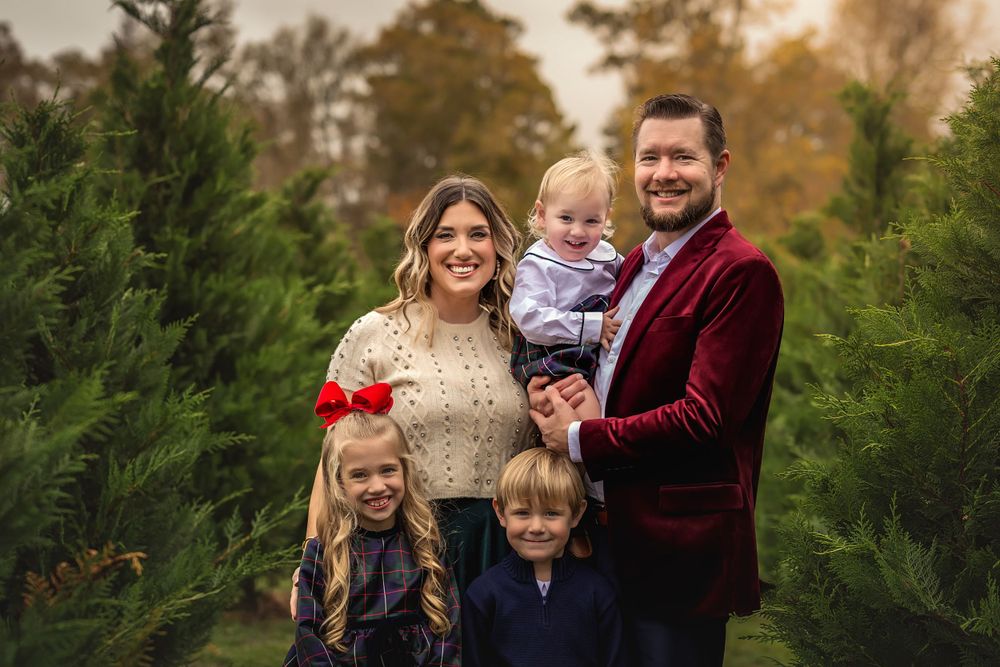 A family poses together at a Christmas tree farm surrounded by evergreen trees at sunset in elegant outfits.