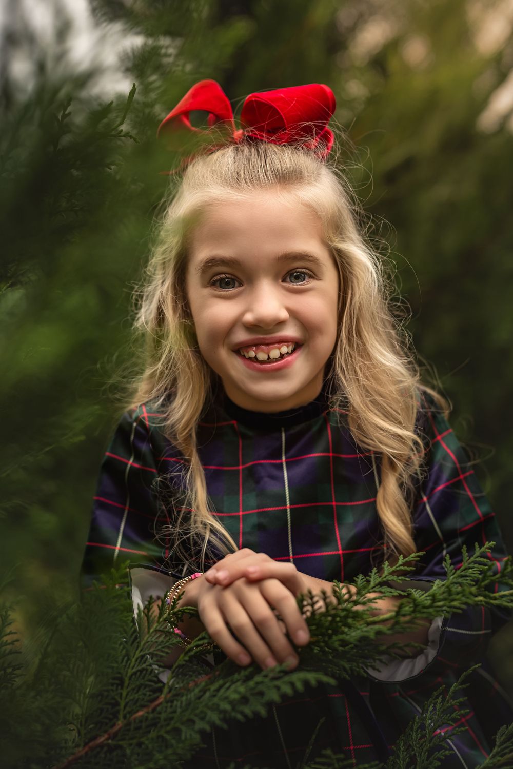A festive holiday portrait of a smiling blonde girl with a plaid dress and red bow amidst evergreen branches.