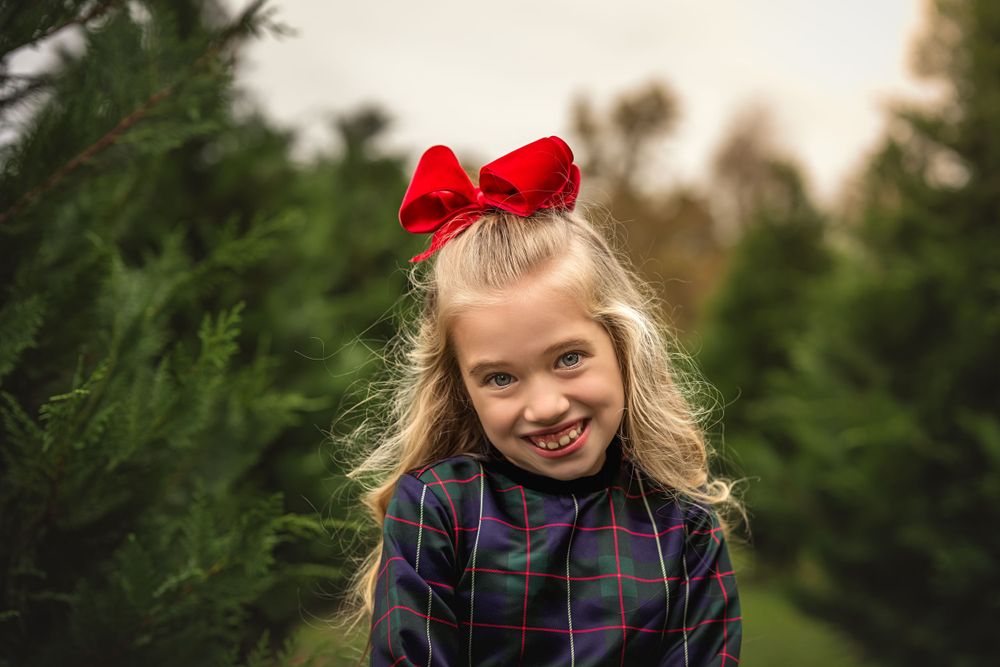 Young child wearing a plaid dress and red bow smiles at camera among evergreen trees at a Christmas tree farm.