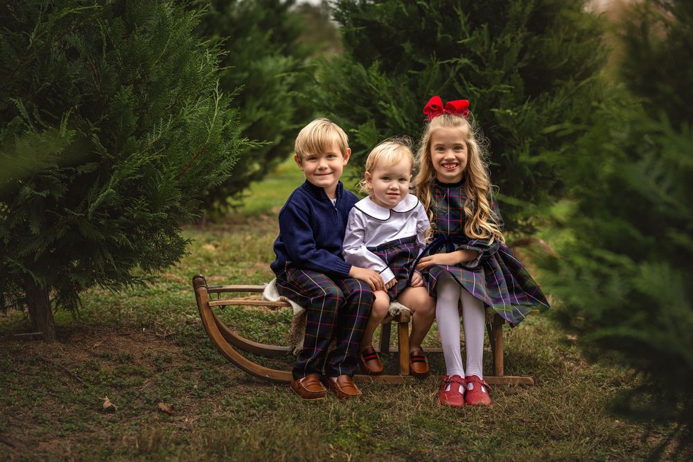 Three children sit together on a vintage wooden sled among evergreen trees during a holiday photoshoot.