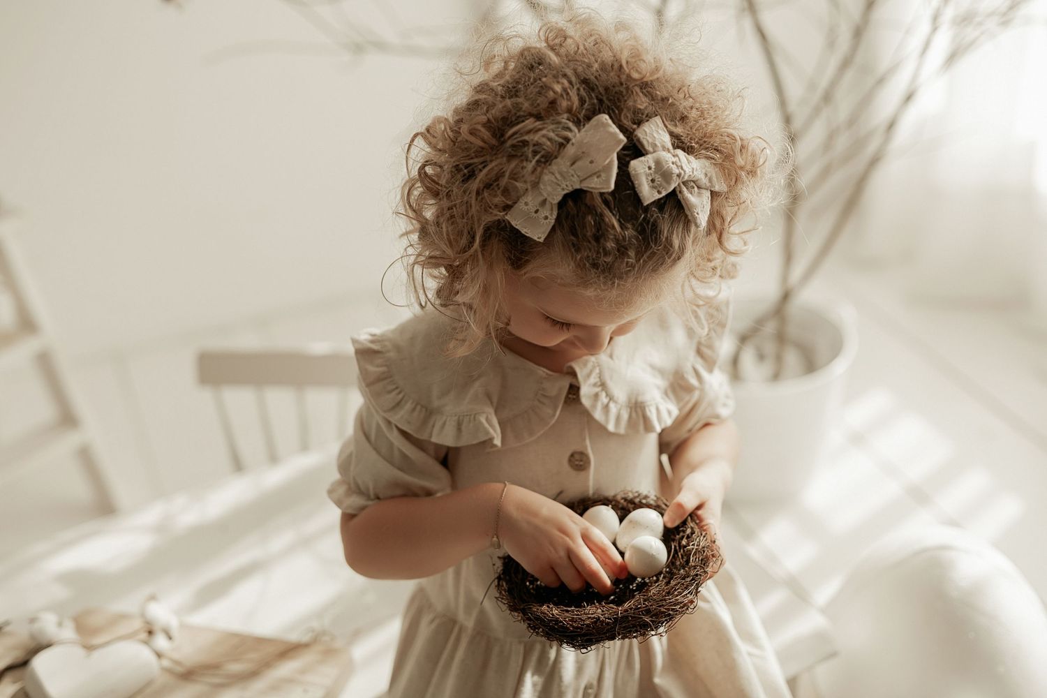 Toddler in vintage-style cream dress with bow headband holds chocolate treats in a dreamy white setting.