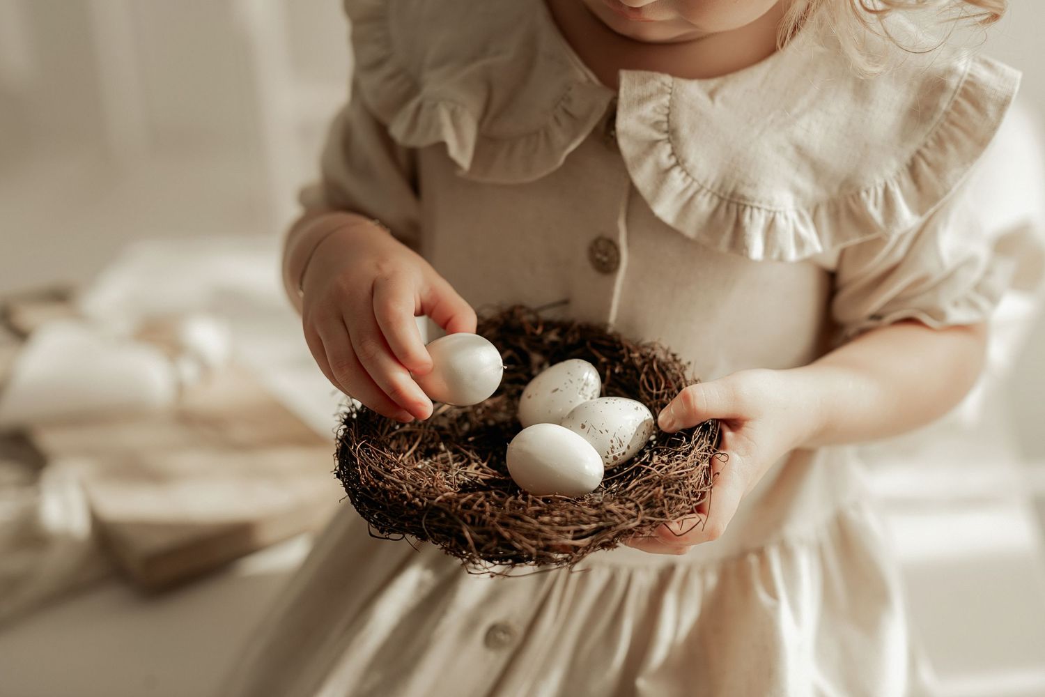 Small hands hold a decorative nest filled with white eggs against a soft neutral background.