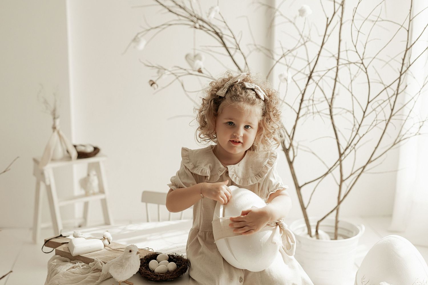 Child in ruffled dress sits among soft white decor with branches and natural elements in dreamy neutral setting.