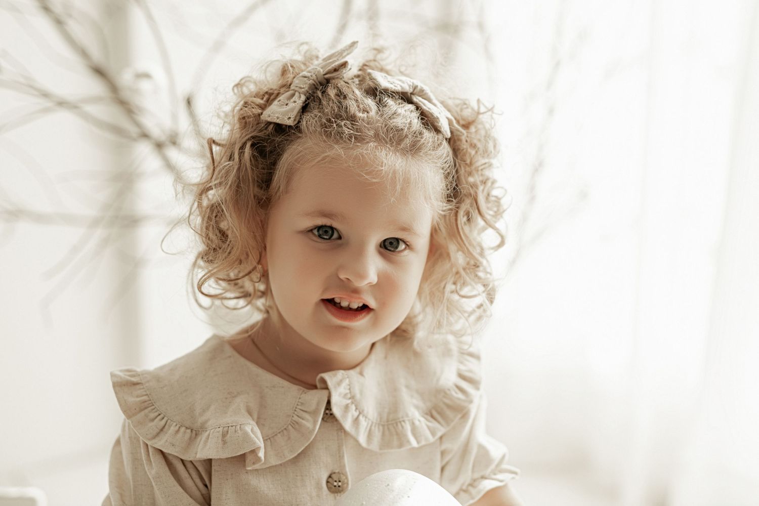 A young child with curly hair wearing a white ruffled dress poses in front of a light backdrop.