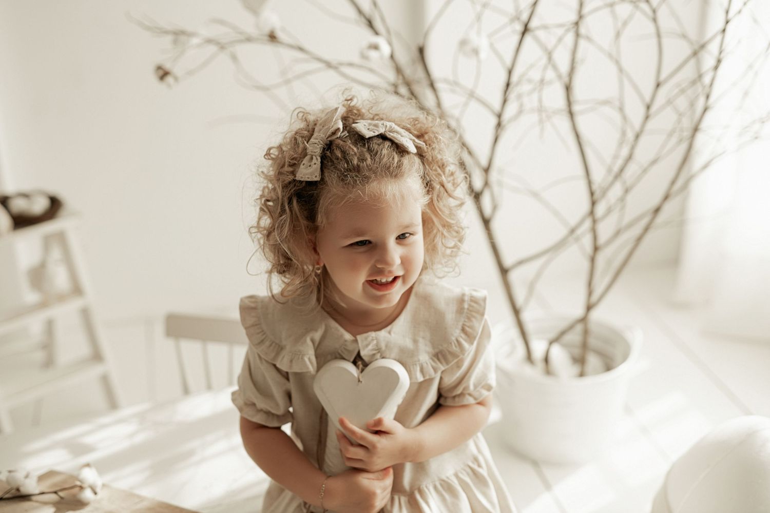 Child in light beige dress holding heart pillow in bright white room with decorative branches.