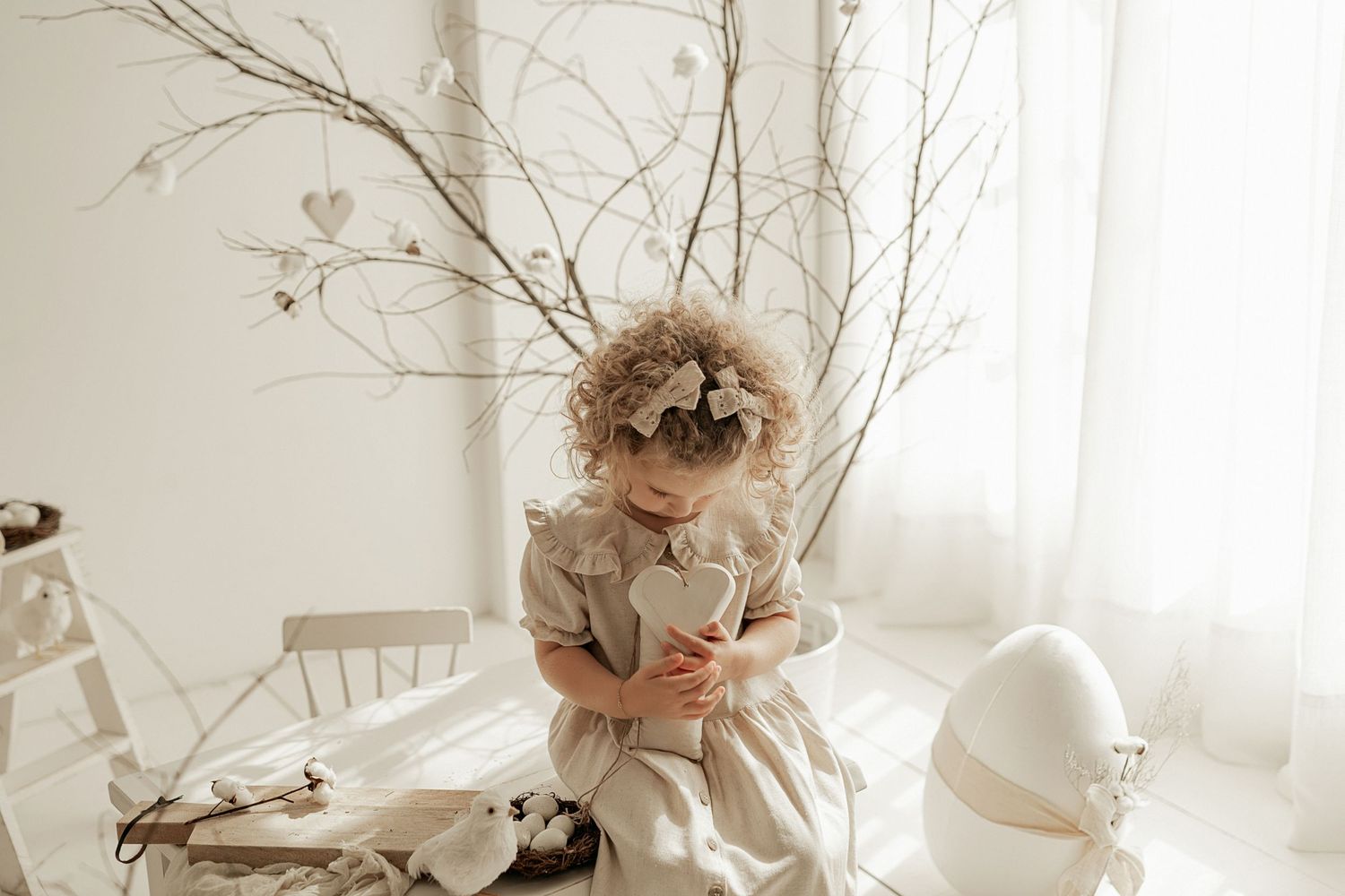 Laughing child sitting by bare branch decor in light neutral room with heart decoration.