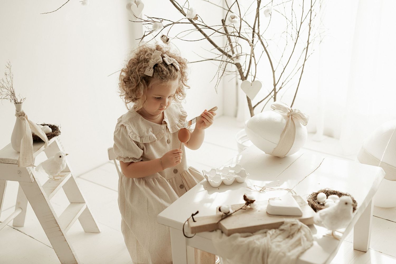 Curly-haired child in white outfit examines items on a white table decorated with bare branches.