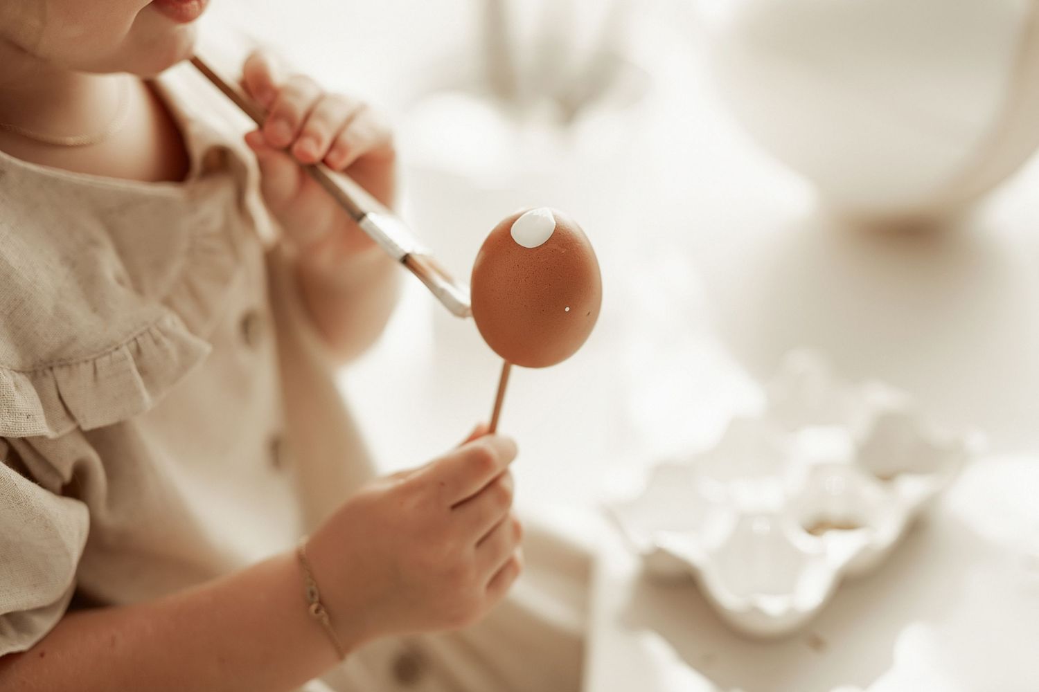 Someone holding a paintbrush delicately painting a brown Easter egg in soft natural light.