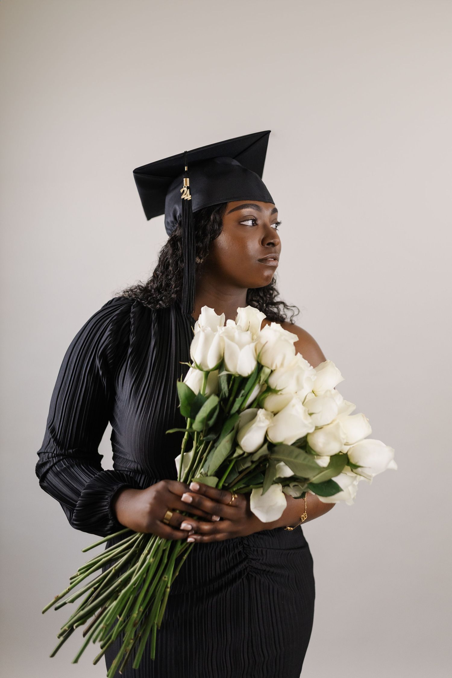 A graduate in cap and gown holds white roses against a neutral background.