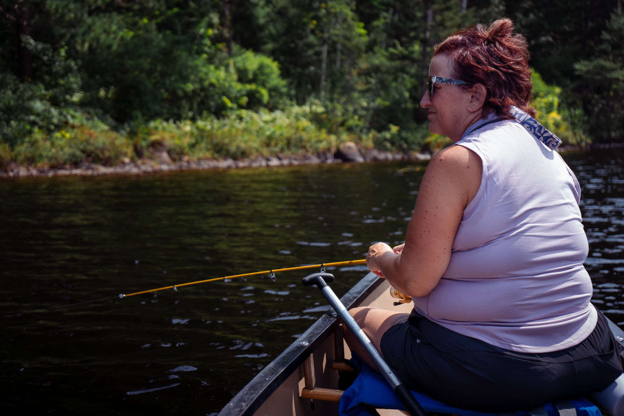 A person in a white tank top sits in a canoe with a fishing pole on a peaceful forest lake.