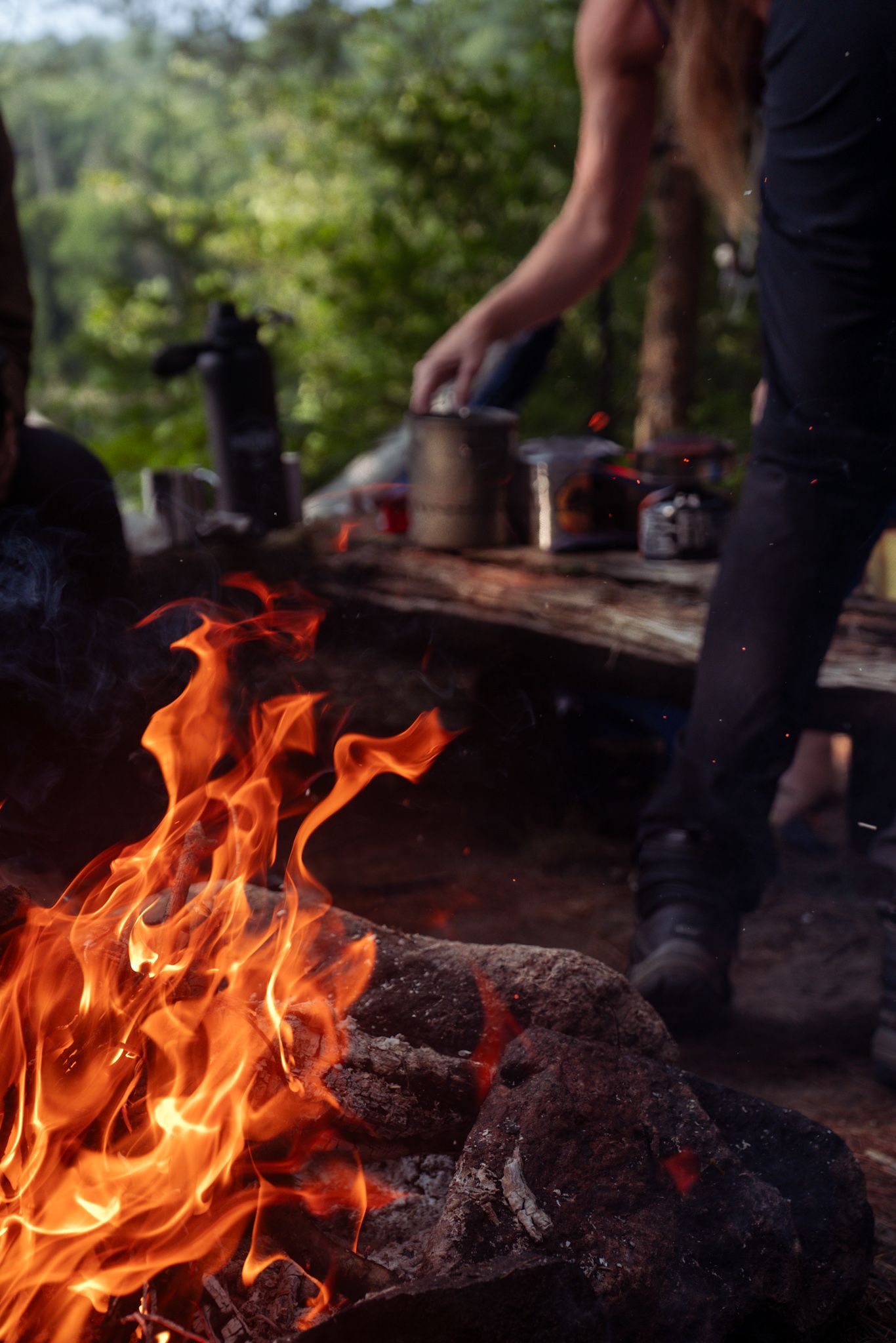 Bright orange campfire flames dance against dark logs while someone cooks outdoors at a rustic camping setup.
