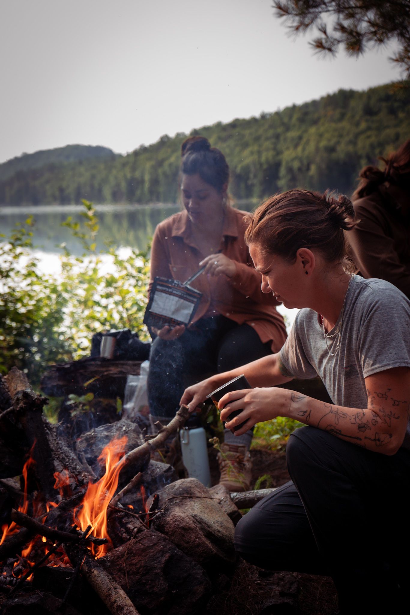 People enjoying a rustic camping experience by a lakeside campfire as sunlight sparkles on the water in the background.