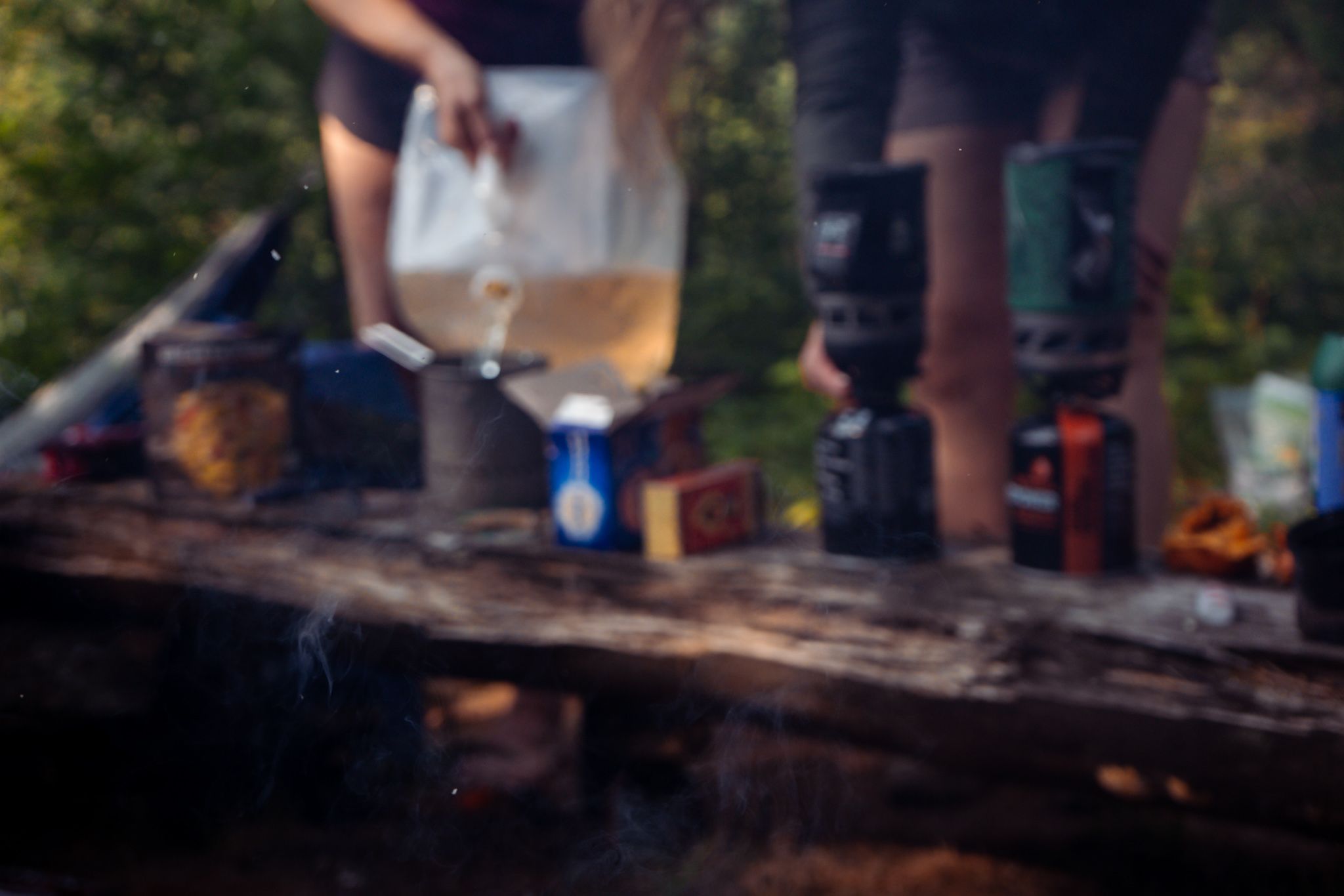 Someone pours a drink from a pitcher at an outdoor gathering with camping equipment on a rustic wooden surface.