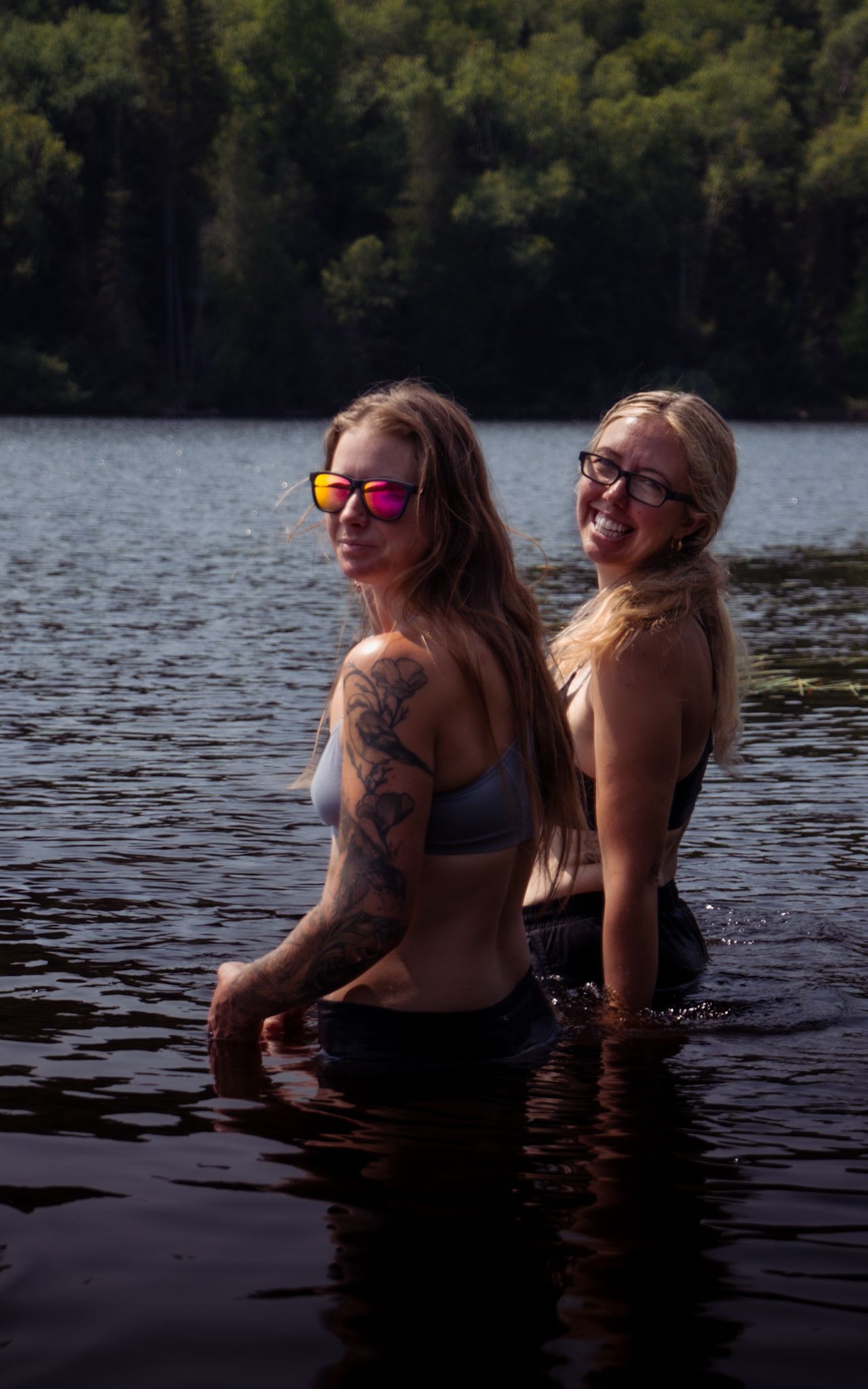 Two women smile while wading in a dark lake surrounded by forest on a summer day.