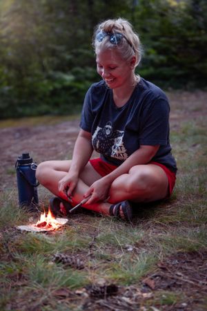 A camper sits cross-legged by a small campfire while wearing a dark t-shirt and red shorts in an outdoor setting.