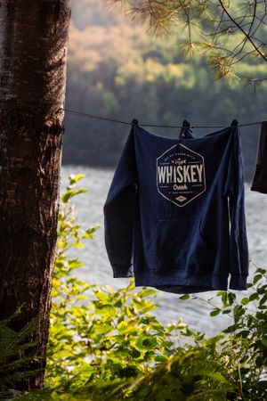 Navy blue t-shirt with whiskey logo hanging to dry on clothesline by lake against sunset backdrop and tree silhouettes.