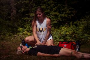 A person performs first aid training on a simulated casualty lying on the ground in a wooded outdoor setting.