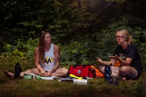 Two people enjoy a summer picnic on a blanket in a wooded area with food and drinks spread out between them.