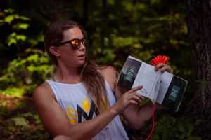 Photographer in white tank top holding camera equipment in a forest setting with dappled sunlight.