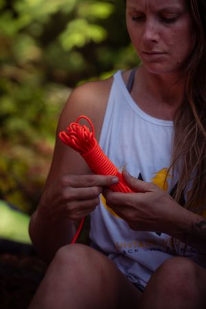 Someone in a white tank top holding red rope or cord in an outdoor setting with blurred greenery in background.