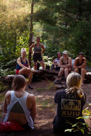 A group of hikers taking a break and resting on logs along a wooded trail during a sunny afternoon.