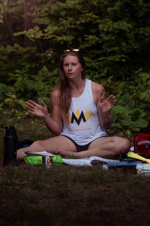 Person wearing white tank top with logo sits cross-legged on ground in wooded setting with water bottle and supplies nearby.