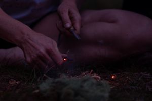Hands light a sparkler in the dark creating glowing embers and smoke against a black background.