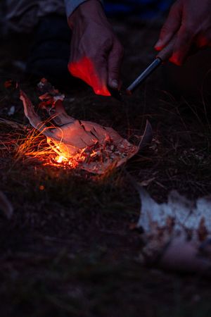 Hands use a ferrocerium rod to create sparks on tinder for fire starting in dark outdoor setting.