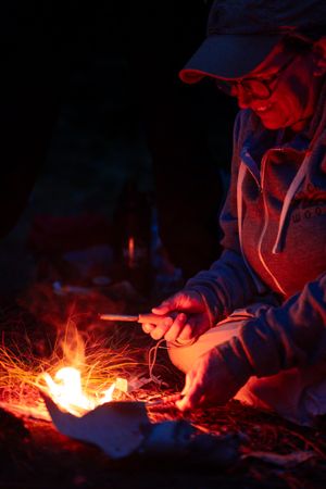 Figure illuminated by campfire light tends to glowing embers against dark night sky with red and blue lighting.