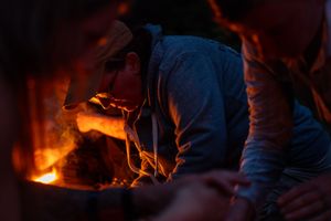 People gather around a glowing campfire at night while tending to the flames in a dark outdoor setting.