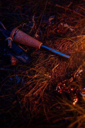 A vintage corkscrew rests in dry grass next to glowing embers in a moody nighttime lighting scene.