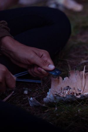 Hands using tongs to tend a small campfire with burning sticks as darkness falls.